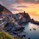 petit-village-cotier-perche-sur-des-rochers-au-coucher-du-soleil-maisons-en-pierre-aux-toits-tuiles-sentiers-sinueux-et-petits-bateaux-amarres-dans-un-port-etroit-mer-calme-et-falaises-en-arr